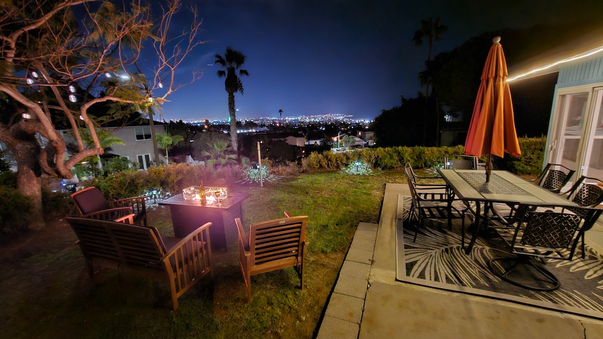 An outdoor space is illuminated at night, featuring a fire pit surrounded by wooden chairs. A six-seat dining table with an umbrella is positioned nearby. The view reveals a city skyline and distant lights, framed by trees and vegetation.