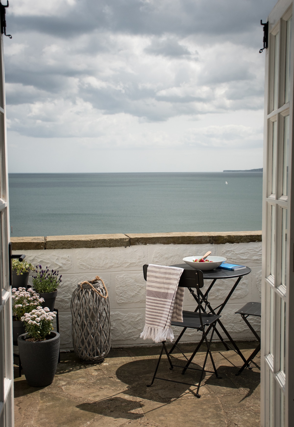 A small balcony is framed by open double doors, revealing sweeping views of the sea. A round black table is set with a white bowl and a striped towel. Potted plants add a touch of greenery next to decorative elements on the stone surface.