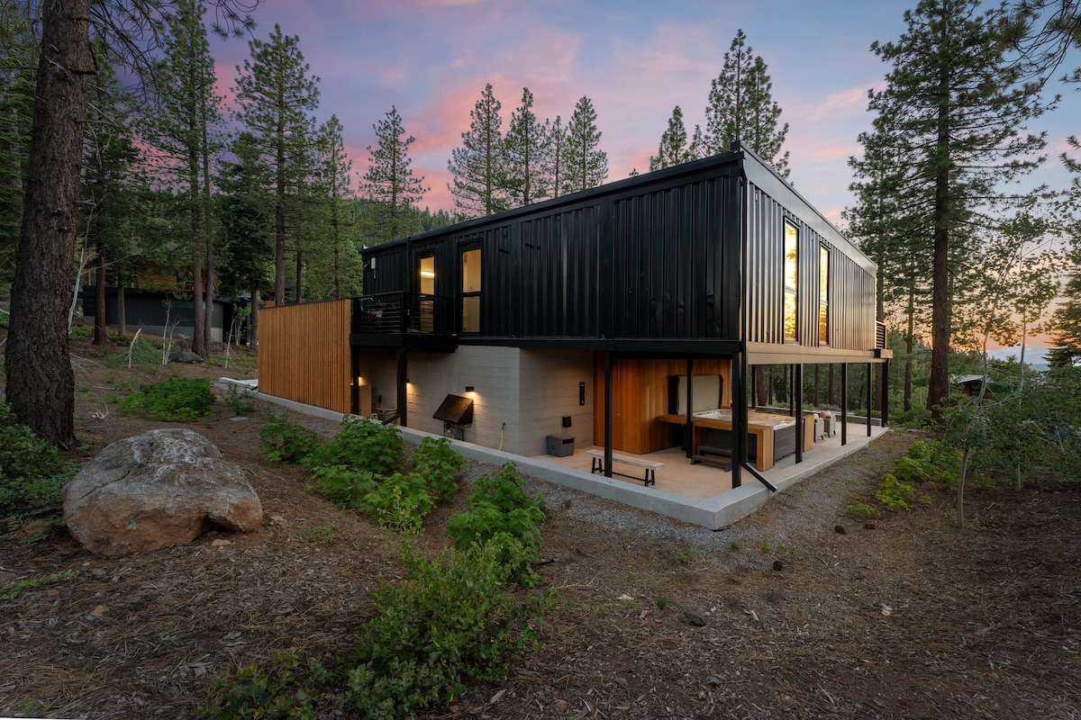 The modern home is set among tall trees, showcasing a sleek black exterior complemented by warm wooden accents. Large windows reflect soft sunset hues, while a spacious outdoor patio area is visible, featuring seating and a landscaped yard with natural elements.