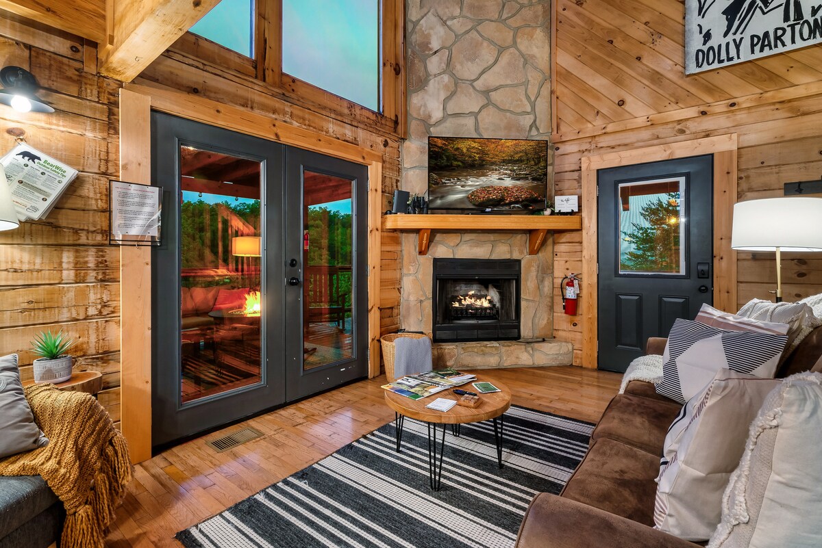 The living room features warm wooden walls and a cozy electric fireplace. Large doors allow natural light and offer views of the outdoors. A 55" Roku Smart TV is mounted above the mantle. A coffee table is situated on a striped rug, accompanied by comfortable seating.