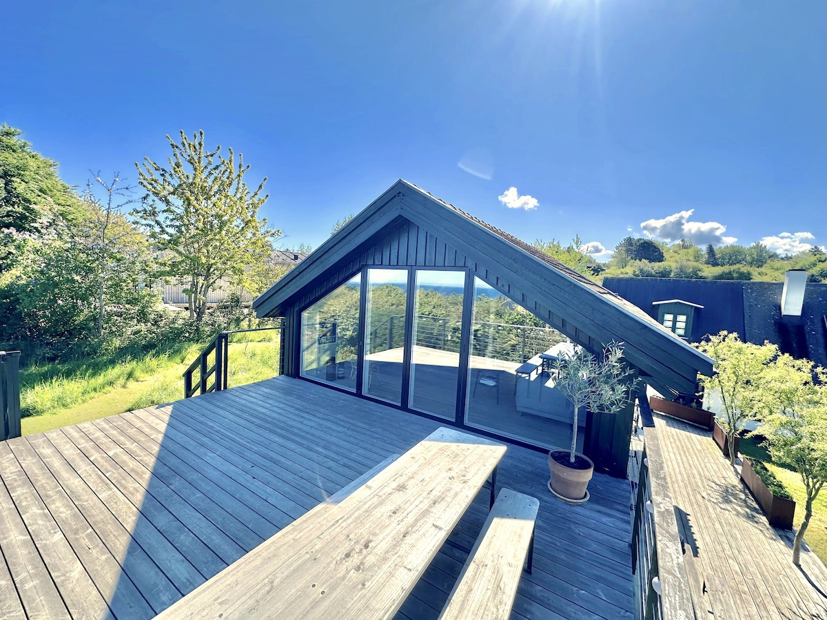 A wooden deck with ample seating is featured, surrounded by lush greenery and open space. A large glass wall offers expansive views of the outdoors, and sunlight fills the area, creating a bright and airy environment.