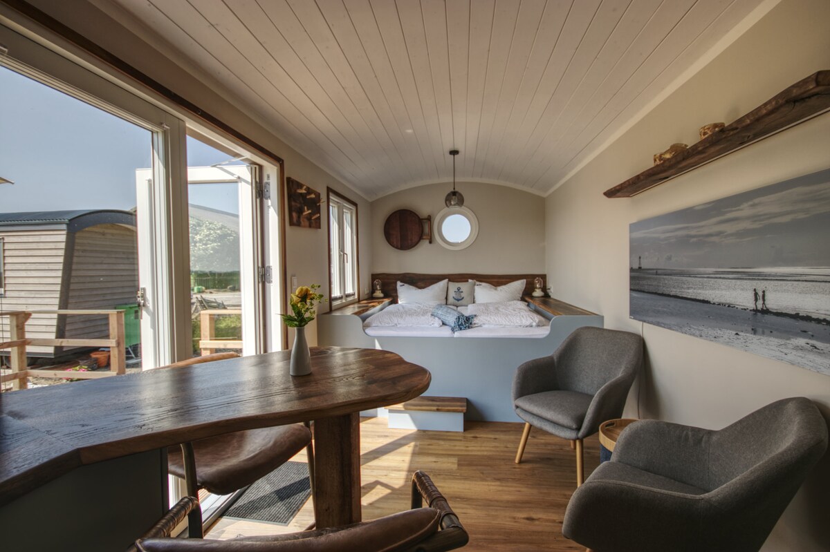 An inviting interior features a cozy sleeping area with a spacious bed adorned with white linens and blue accents. A small dining table is positioned by large windows, allowing natural light to fill the space. Two armchairs complement the rustic wooden decor.