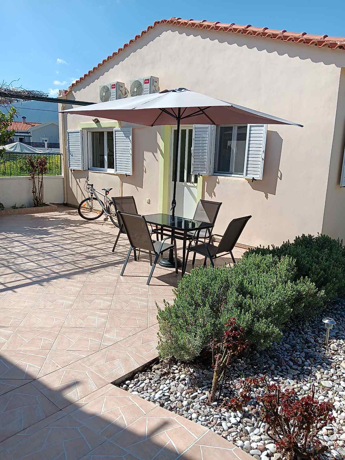 A patio area features a black metal table surrounded by four chairs, all shaded by a large umbrella. Decorative stones and low shrubs add greenery alongside the tiled floor. A bicycle leans against the wall of the house, which has two windows with shutters.