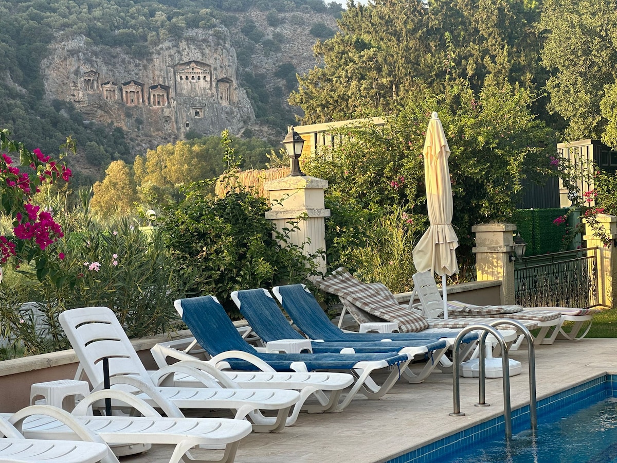 A serene outdoor area features several lounge chairs positioned beside a swimming pool. Vibrant flowers and lush greenery surround the space, enhancing the view of the historical Caunos King Tombs carved into the hillside in the background.