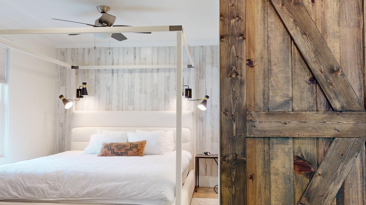 A spacious bedroom features a modern four-poster bed with ample white bedding, illuminated by stylish wall sconces. A textured wooden wall adds a rustic touch, complemented by a sleek ceiling fan. The image also highlights a closed wooden door with a classic barn-style design.