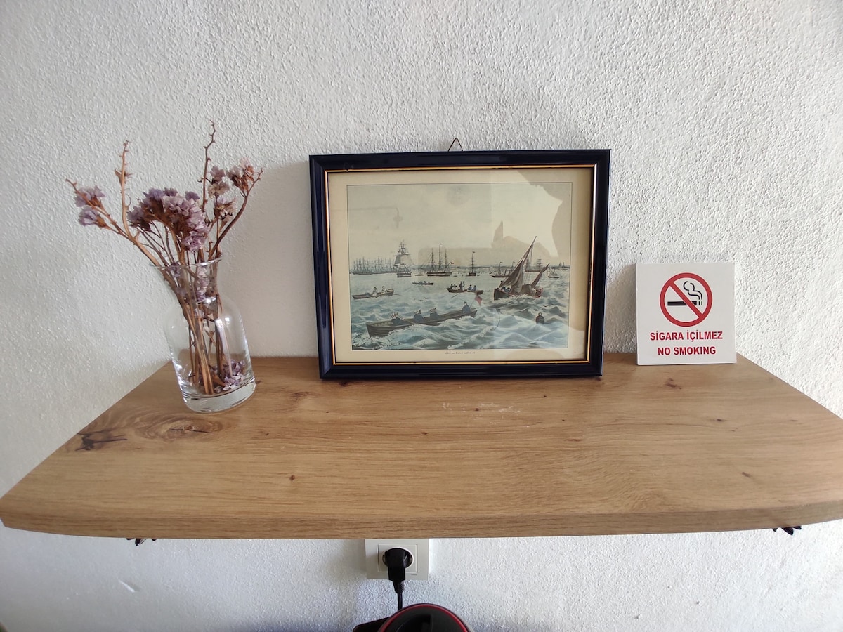 A wooden shelf displays a framed painting of a maritime scene, depicting boats on turbulent waters. A small vase holds dried flowers, and a sign reading 'No Smoking' is positioned beside the artwork, highlighting the room's decor and adherence to smoke-free guidelines.