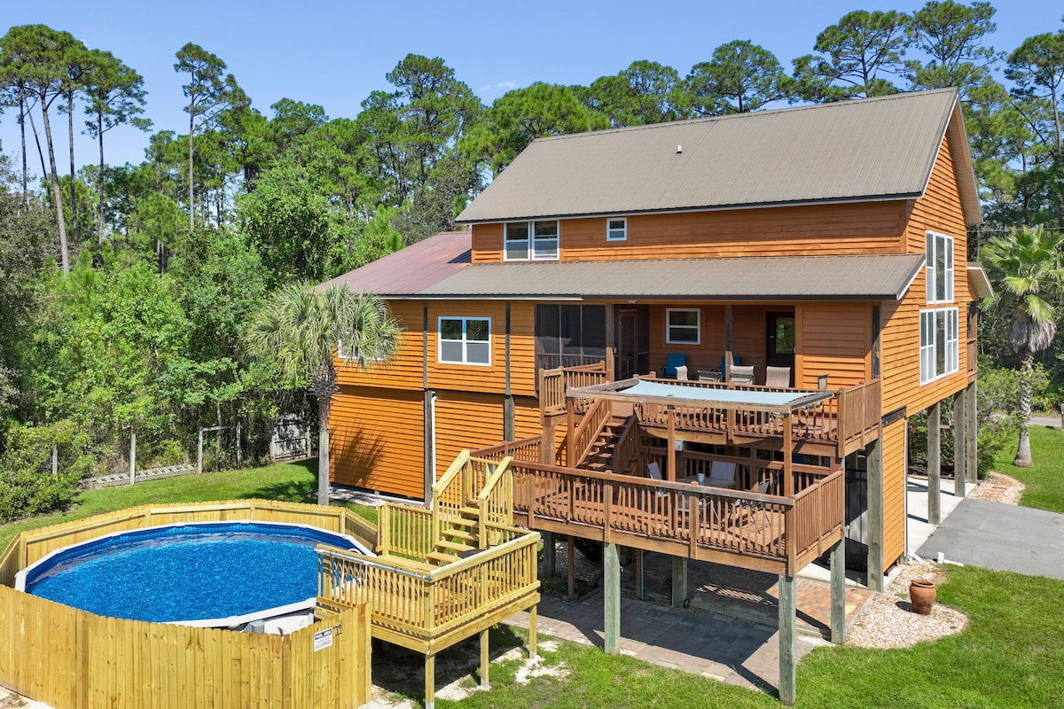 A large orange house features multiple decks that overlook a circular swimming pool surrounded by wooden decking. Lush greenery and tall trees provide a natural backdrop, enhancing the outdoor space for relaxation. Ample seating can be seen on the upper deck.