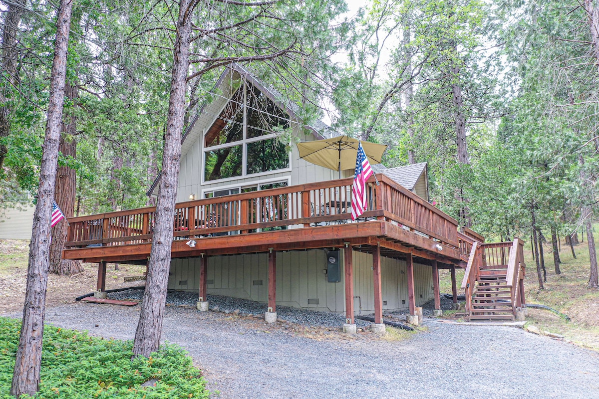 The cabin's exterior showcases a spacious deck with seating, surrounded by tall trees. Large windows reflect natural light, enhancing the A-frame design. American flags adorn the railing, and a staircase leads from the deck to the gravel driveway.