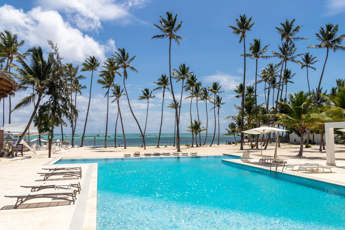 A large swimming pool is situated in front of a sandy beach, surrounded by palm trees swaying gently in the breeze. The clear blue water of the pool contrasts with the ocean in the background, creating a serene atmosphere under a bright sky.