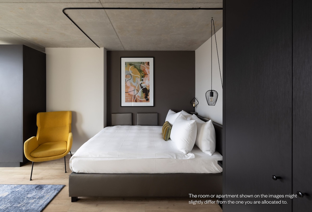 A spacious bedroom is depicted, featuring a comfortable double bed with white bedding and plush pillows. A bright yellow accent chair stands nearby, complemented by a contemporary wall art piece. Neutral tones and wooden flooring enhance the room's overall modern aesthetic.