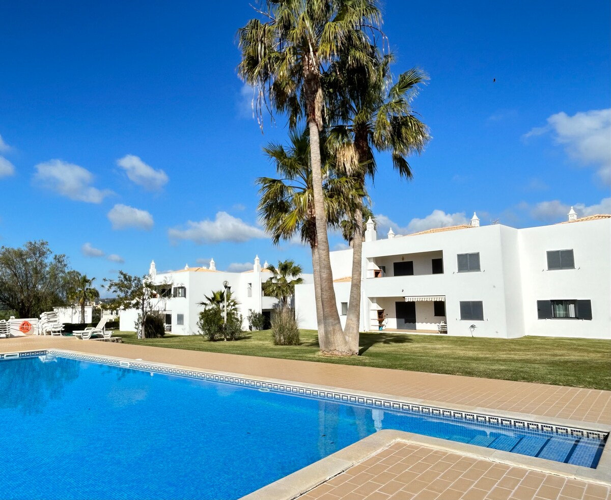 A clear blue swimming pool reflects the sunny sky, framed by neatly trimmed grass. Nearby, palm trees provide shade, while two white buildings with balconies are visible in the background, set amidst a peaceful, landscaped area.