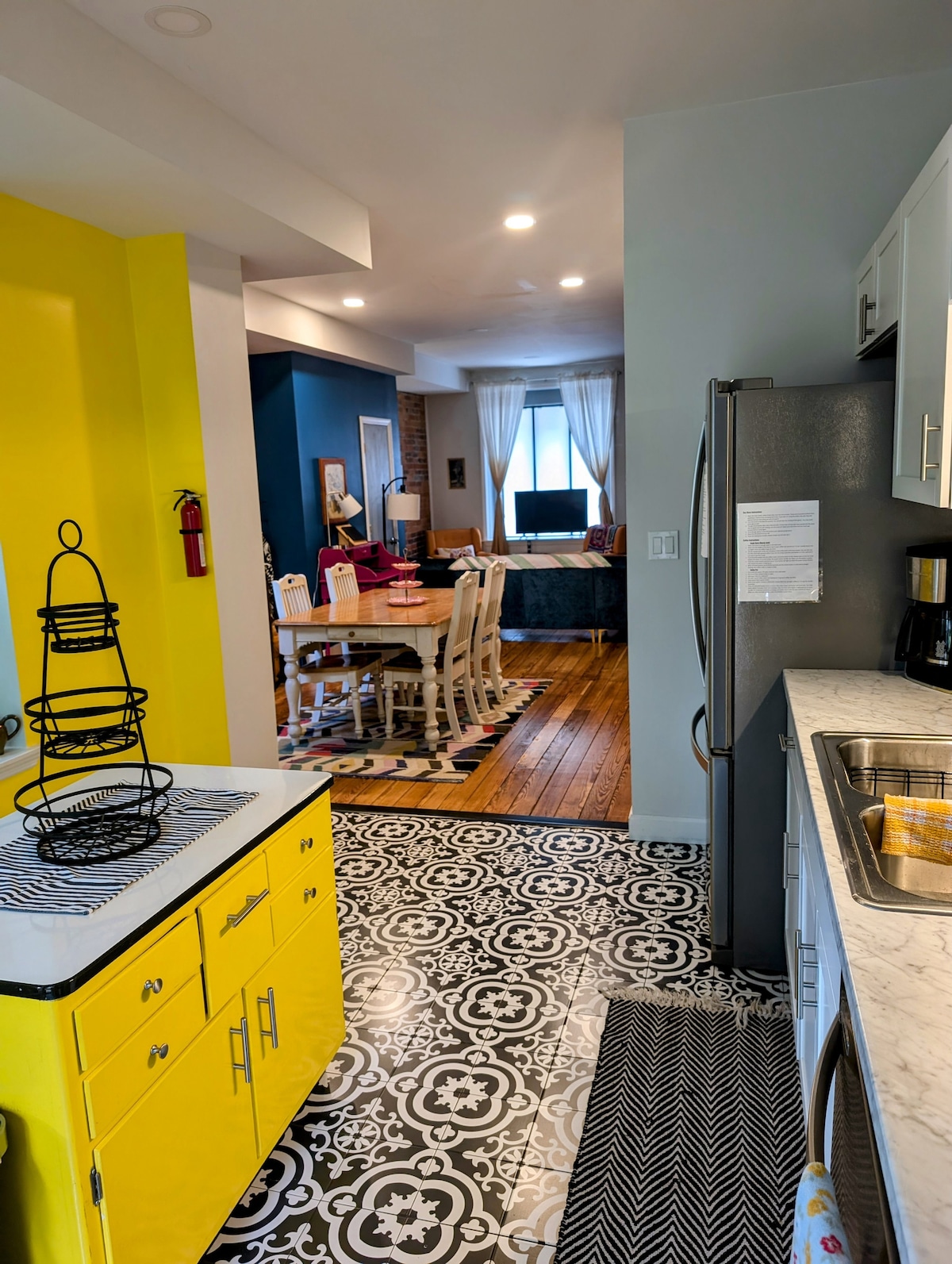A modern kitchen is visible, featuring bright yellow cabinetry and a patterned black and white floor. To the left, an open layout leads into a dining area with a wooden table and multiple chairs, illuminated by natural light from a nearby window.