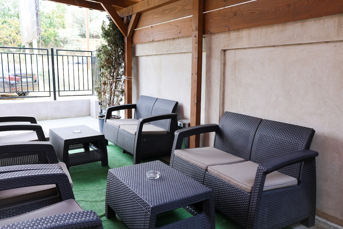 A shaded outdoor seating area features several cushioned chairs and a low coffee table, arranged on green artificial grass. The wooden structure provides shelter, while nearby greenery adds a touch of nature to the space.