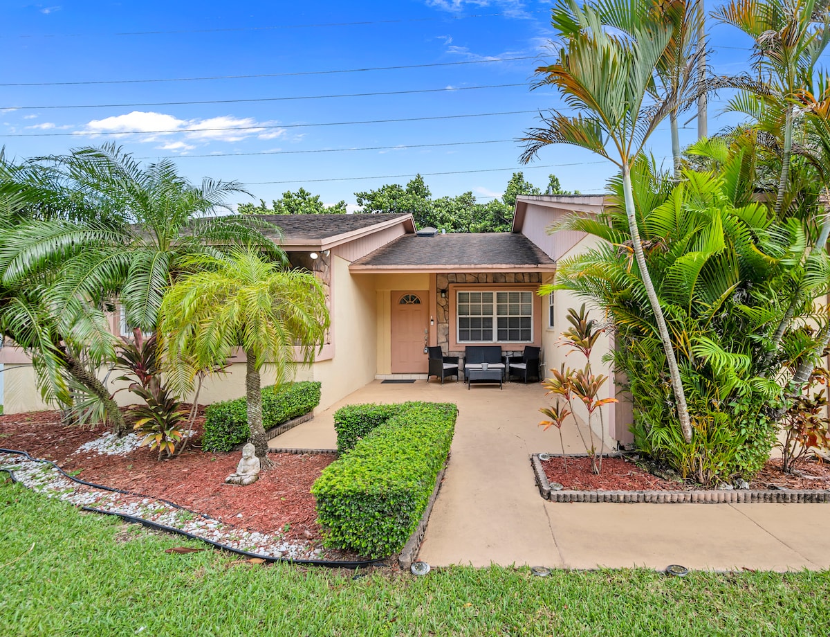 The front entrance is framed by lush tropical landscaping and palm trees. A small seating area with dark chairs provides a welcoming touch. A path leads to the entrance of the home, enhancing the inviting atmosphere of the property.