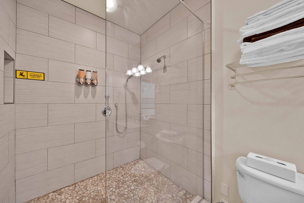 A spacious walk-in shower is enclosed by glass panels, featuring natural stone flooring. Shower fixtures are positioned along the wall, illuminating the area with soft lighting. Clean towels are neatly arranged on a shelf above, while a toilet is visible in the corner.