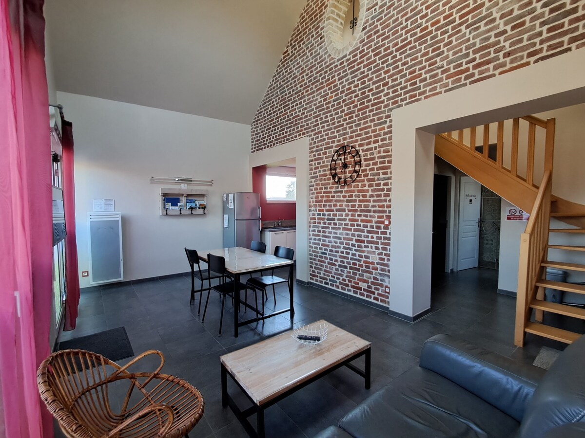 An open living area features exposed brick walls and high ceilings. A spacious dining table is set for four, while a comfortable seating arrangement includes a dark sofa. A staircase leads to the upper level, with a modern kitchen visible in the background, equipped with stainless steel appliances.