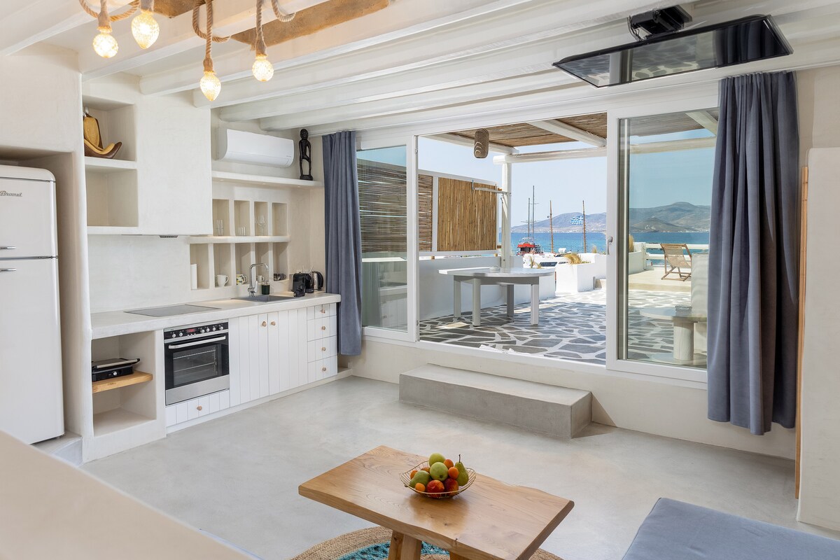 A bright and airy living area features a modern kitchen setup with essential appliances and a dining space. Large glass doors provide an unobstructed view of the outdoor area and sea. Natural light fills the space, accentuating the minimalist design.