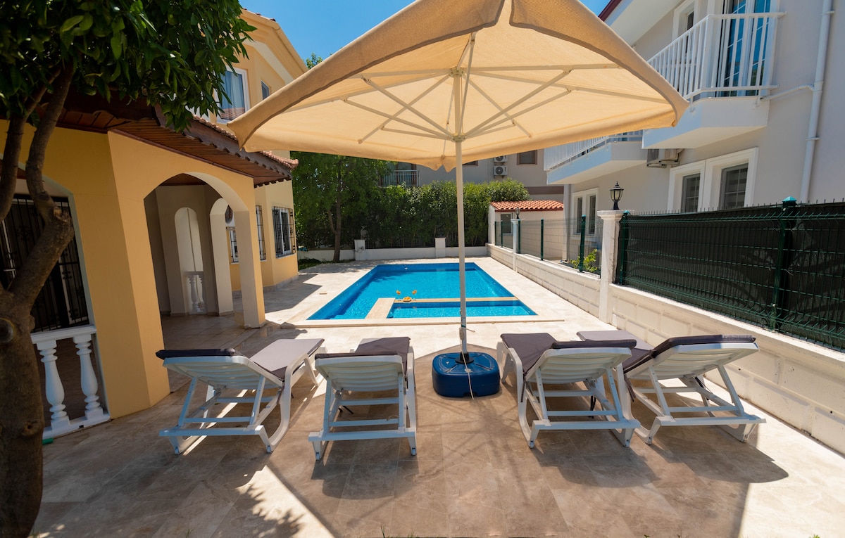 A well-maintained outdoor area features a private swimming pool surrounded by a sun terrace. Four sun loungers are arranged under a large umbrella, providing shade. The villa's colorful exterior can be seen alongside lush greenery, creating a tranquil environment.