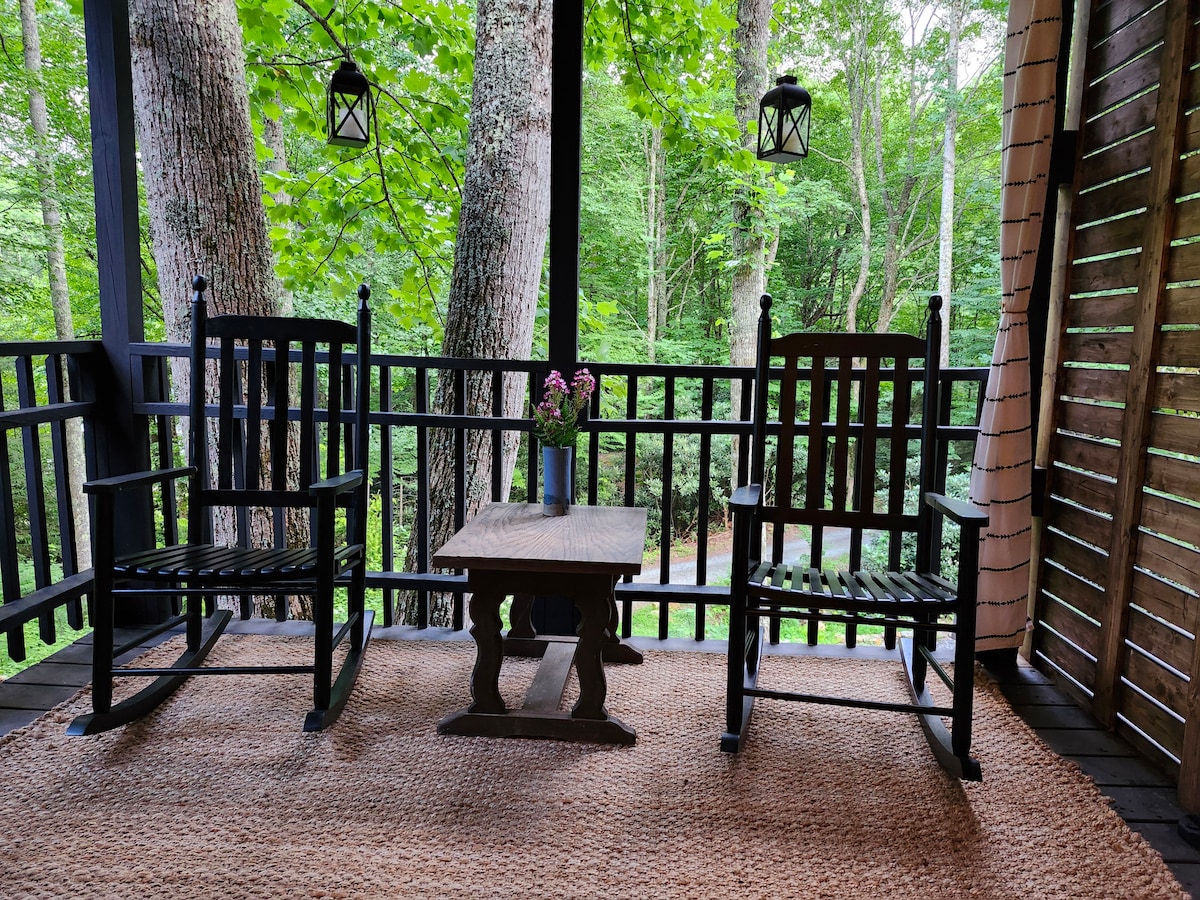 A cozy outdoor seating area features two black rocking chairs positioned around a rustic wooden table. A small vase with flowers sits on the table, surrounded by a woven rug. Tall trees create a natural backdrop, enhancing the tranquil woodland setting.