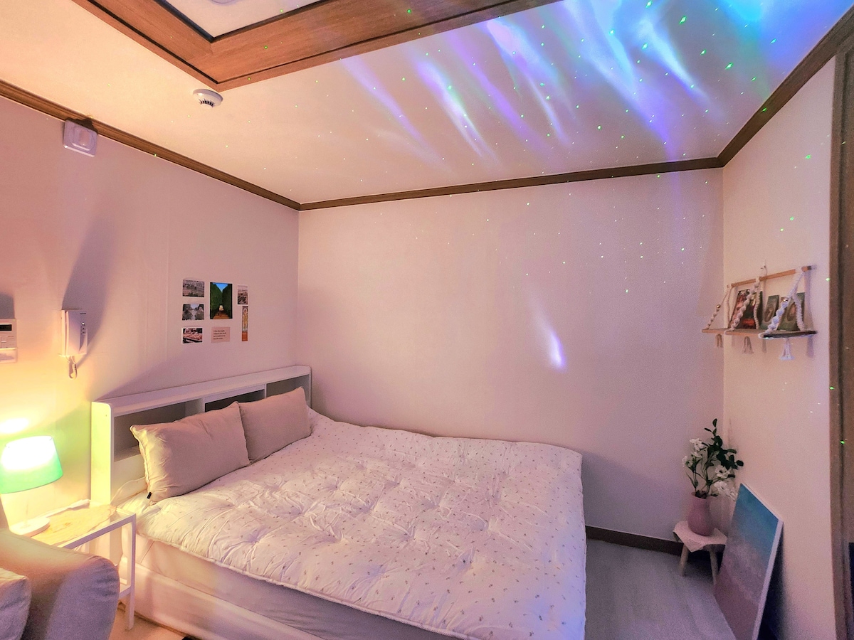 A spacious sleeping area features a large bed covered with a textured, light-colored blanket. Subtle star-like lights are projected onto the walls and ceiling, creating a calming ambiance. Decorative items are neatly arranged on a shelf, and a small plant adds a touch of greenery.
