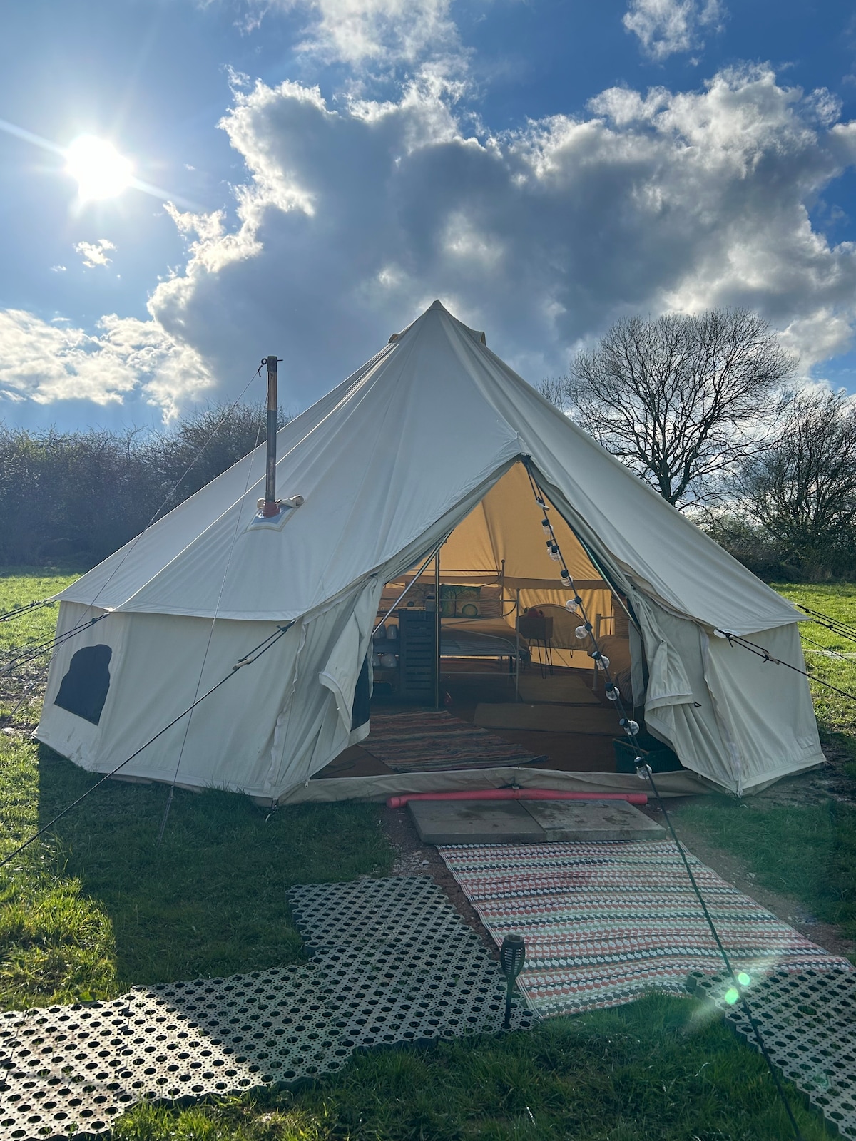 A spacious bell tent is positioned in a grassy area, with a clear entrance showcasing its interior. The structure features a wood-burning stove visible inside, while natural light filters through. The scene is set under a bright sky with scattered clouds.