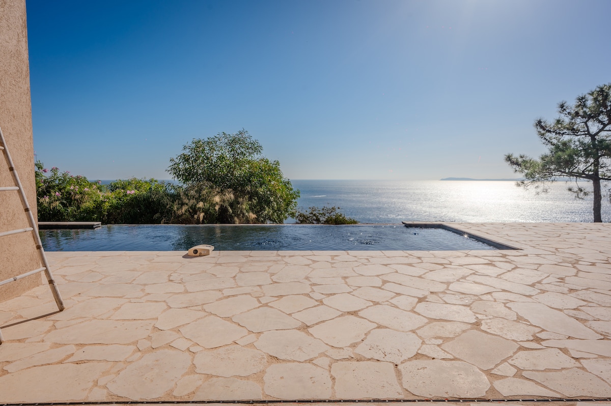 A serene infinity pool overlooks the ocean, with a smooth stone terrace extending from it. Lush greenery frames the scene, enhancing the views. The clear sky and calm waters create a tranquil atmosphere, inviting relaxation and enjoyment of the surrounding natural beauty.
