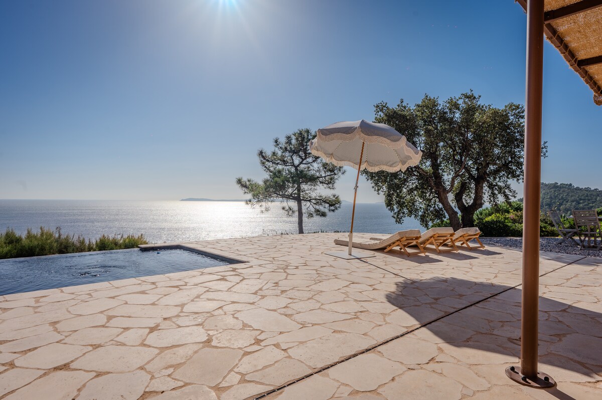 A spacious outdoor terrace offers sun loungers and a large umbrella, overlooking a tranquil infinity pool that reflects the sunlight. The panoramic view of the sparkling sea and distant island can be seen in the background, complemented by lush greenery and trees.