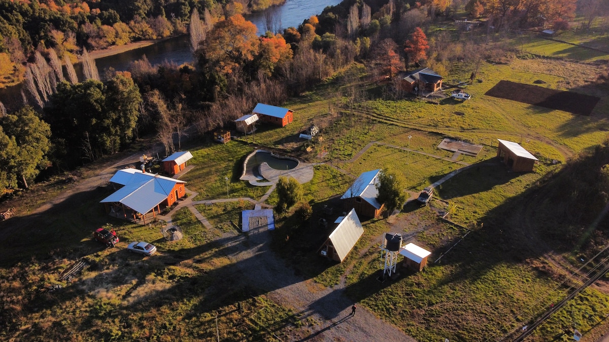 An aerial view showcases the rustic landscape of the cabin complex, featuring multiple orange-hued cabins amidst lush greenery. A meandering river complements the scenery, while a swimming pool is nestled within the grounds. The sunlight casts gentle shadows across the peaceful surroundings.