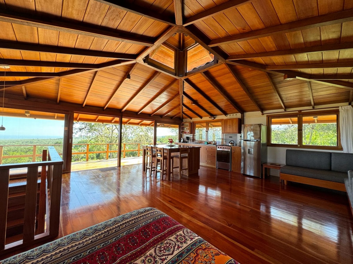 An open-plan living area displays rich wooden beams and a polished hardwood floor. A spacious kitchen with stainless steel appliances is seen alongside a dining table. Large windows provide sweeping views of the surrounding landscape, enhancing the connection with nature.