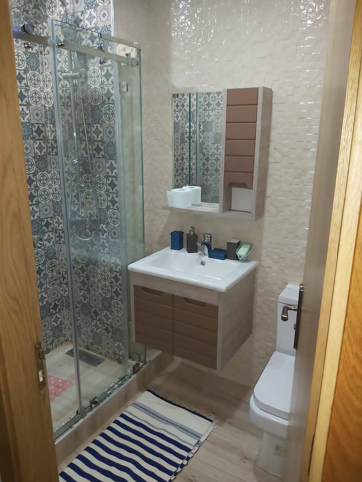 A modern bathroom features a walk-in shower with sliding glass doors, complemented by patterned tiles. A sleek vanity with wooden drawers provides ample storage, while a mirror above reflects natural light. The space is completed with a toilet and a striped rug for added comfort.