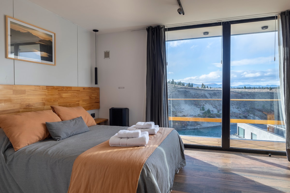 A spacious sleeping area features a wooden headboard and neutral-coloured bedding. Large glass doors open to a scenic view of the landscape, allowing natural light to fill the room. Towels are neatly arranged on the bed, and dark curtains frame the glass doors.