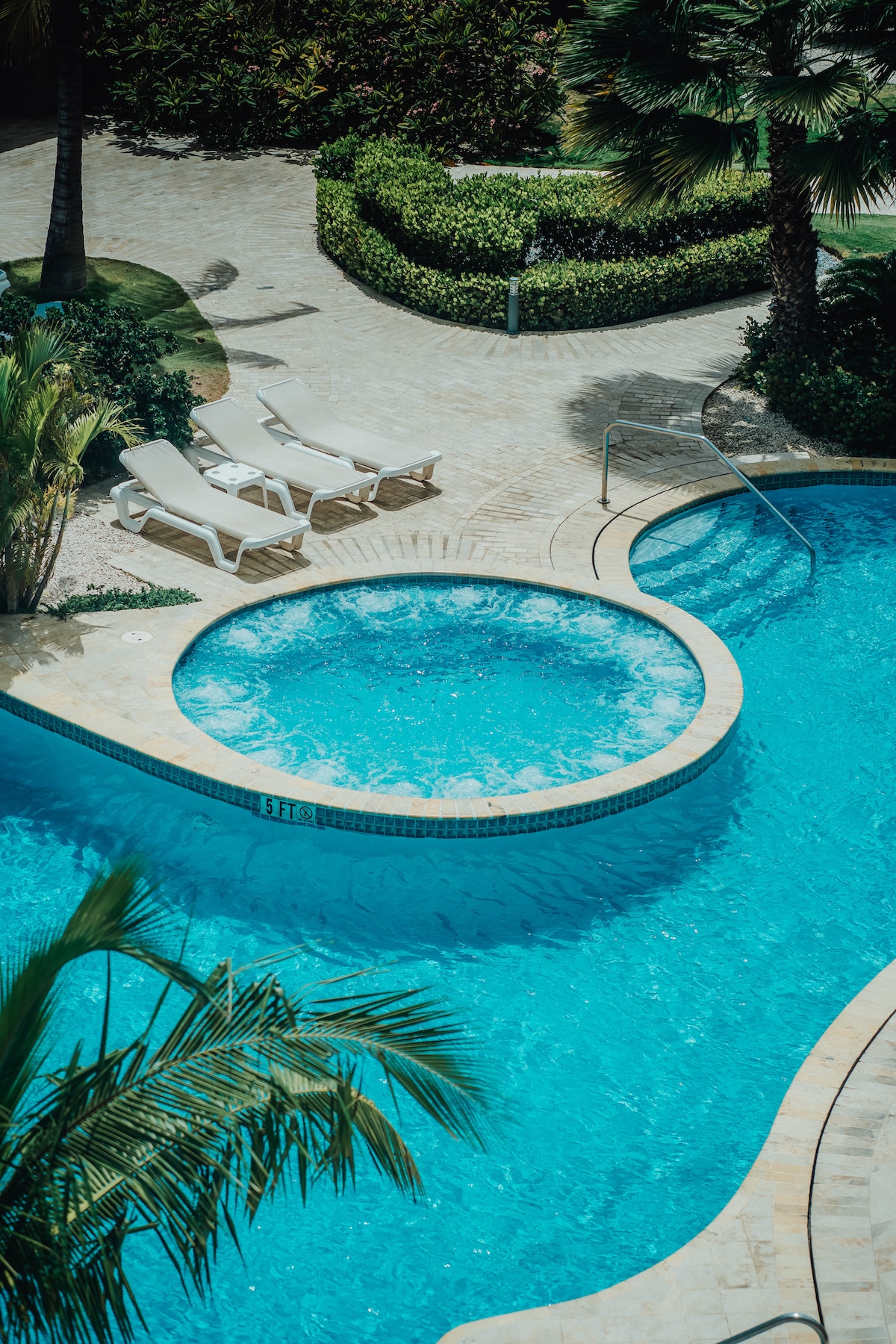 A swimming pool area featuring a large main pool and a circular jacuzzi, both surrounded by lush greenery. Two lounge chairs are positioned nearby, providing a tranquil space for relaxation. The smooth stone pathways integrate with the natural landscaping.