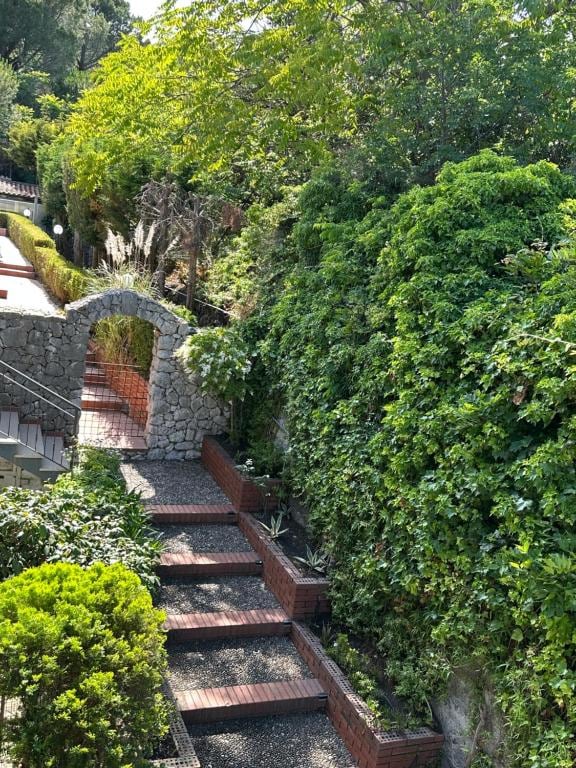 A serene outdoor pathway is lined with lush greenery, featuring a stone archway leading to a charming garden. Brick steps and decorative plants create a peaceful ambiance, inviting visitors to explore the surrounding natural beauty.