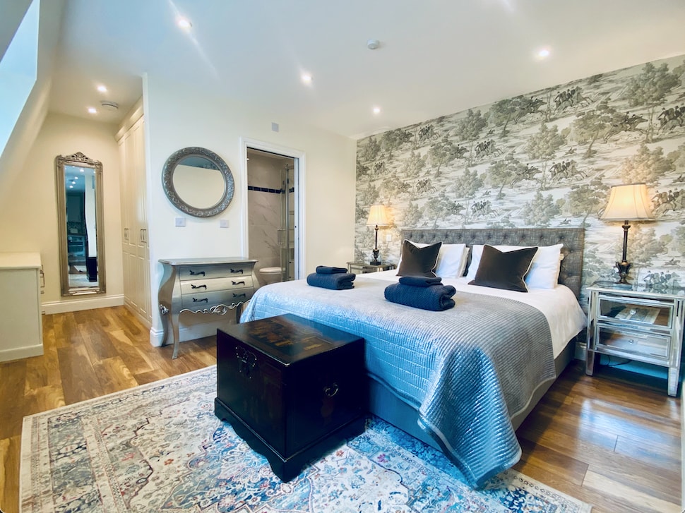 A well-appointed bedroom features a spacious bed with quality linens and decorative throw pillows, complemented by bedside tables and elegant lamps. A vintage-style mirror and cabinet add character, while the wallpaper enhances the room's charm. Warm wood flooring completes the inviting atmosphere.