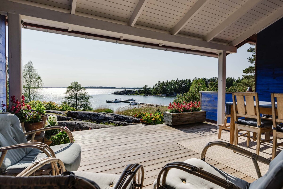 A spacious outdoor patio area features comfortable seating and a wooden table, overlooking serene water and lush greenery. The unobstructed view reveals nearby boats and a calm sea, with colorful flowers adding a natural touch to the setting.