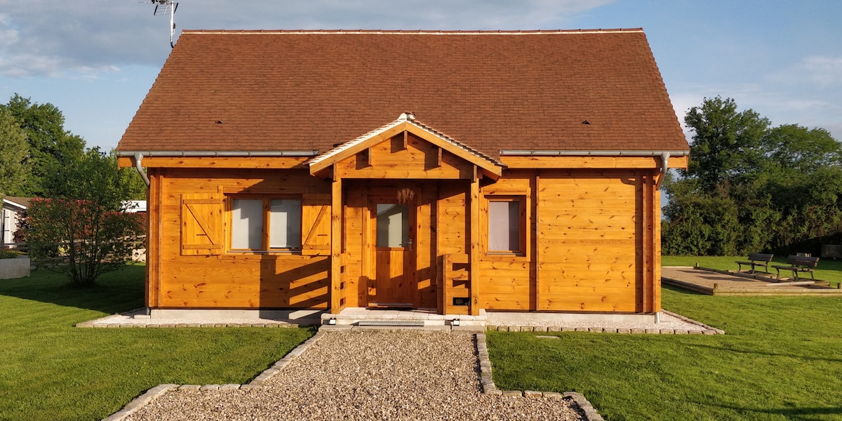 A wooden chalet with a sloped roof is situated in a grassy area. The front entrance features a porch and double doors framed by two windows. Surrounding greenery enhances the peaceful setting, with a gravel path leading to the front door.