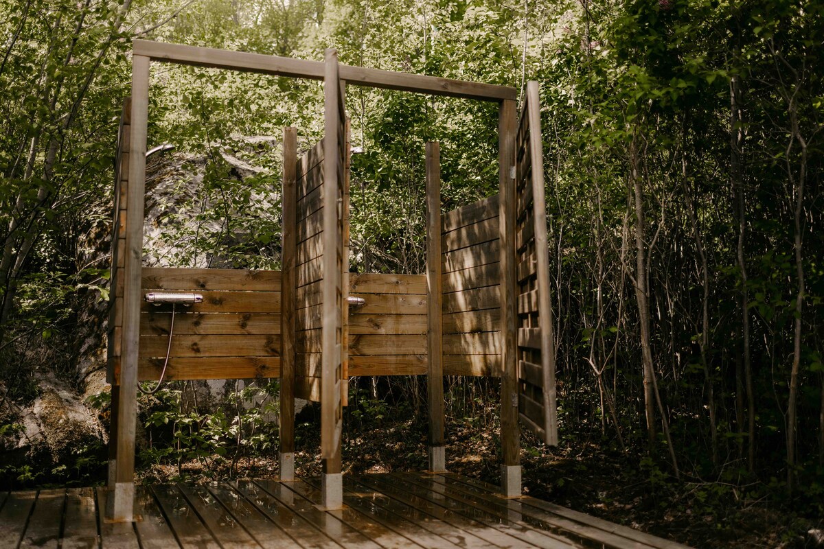 An outdoor shower area is constructed with wooden panels, providing privacy. Surrounded by lush greenery, the space features two shower heads mounted on wooden frames. A wooden deck supports the structure, blending harmoniously with the natural environment.