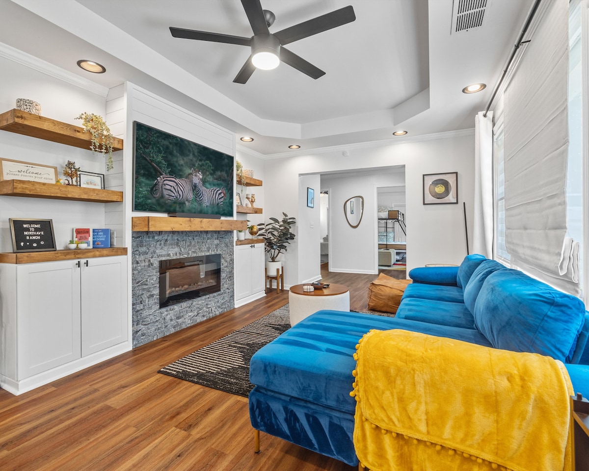 The modern living area features a plush blue sectional sofa complemented by a bright yellow throw blanket. A sleek electric fireplace is positioned beneath a large flat-screen TV displaying wildlife. Shelves with decorative items and plants add character to the space, illuminated by overhead lighting.