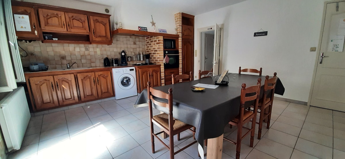The kitchen area features wooden cabinetry and countertop appliances, including a gas stove, oven, and refrigerator. A washing machine is also present. A large table with six chairs occupies the center of the space, complementing the tiled flooring and neutral-colored walls.