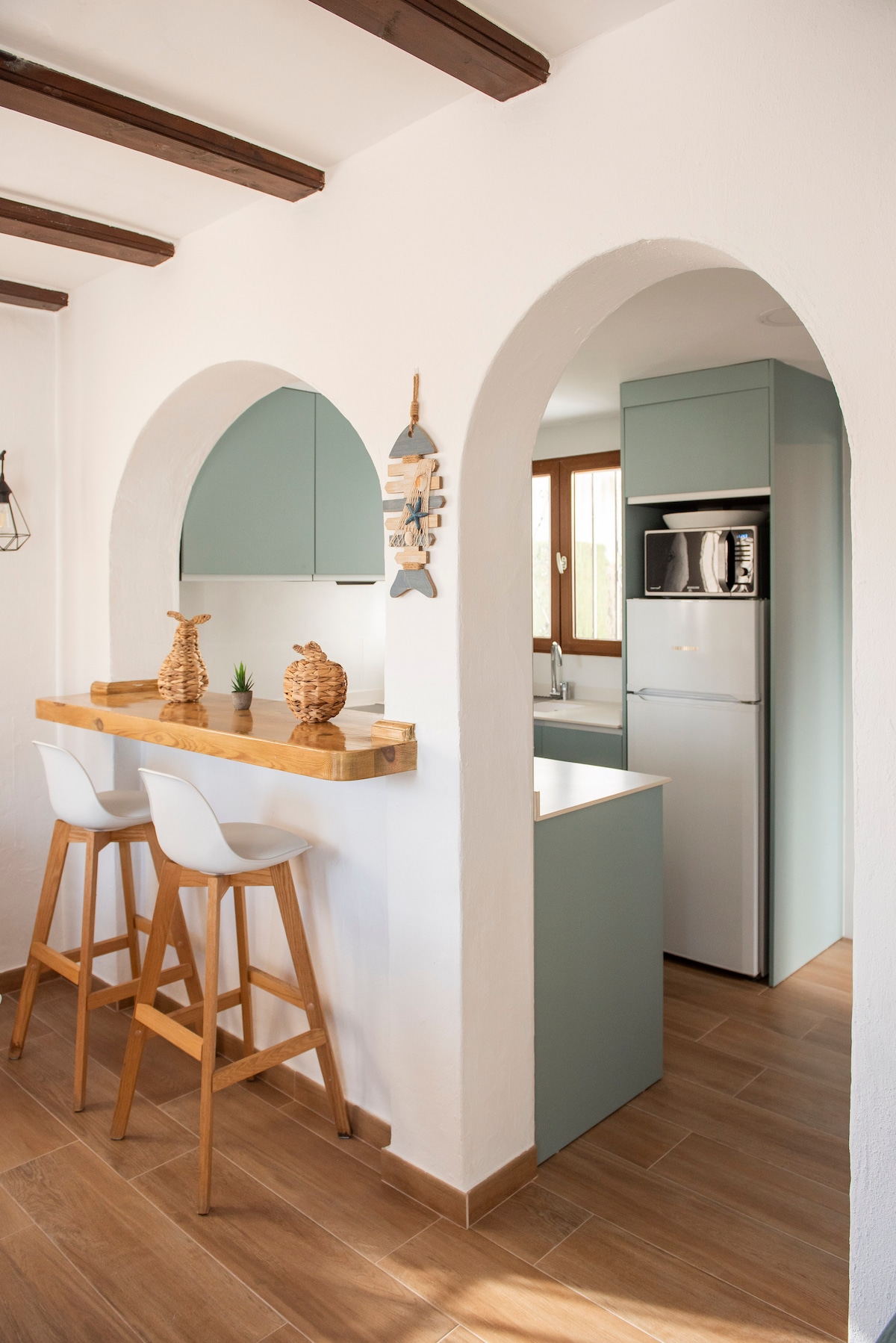 A functional kitchen area features light wooden beams on the ceiling and a bar for two with tall wooden stools. The kitchen includes a fridge and modern cabinetry in soft green tones, complemented by natural light streaming through large windows.