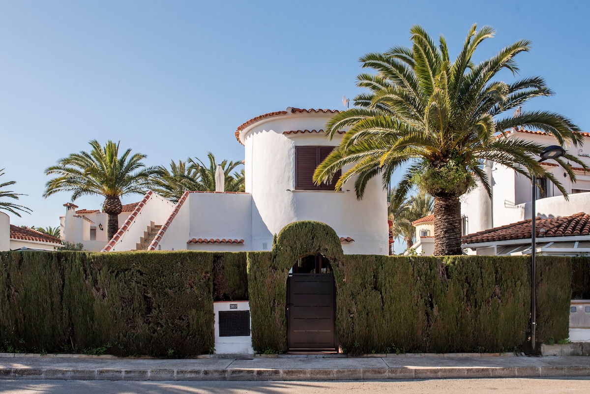 A welcoming entrance is framed by neatly trimmed hedges and palm trees, providing privacy and a tropical ambiance. The white, two-story building features a round tower and traditional tiled roof, highlighting the charming architectural style of the property.