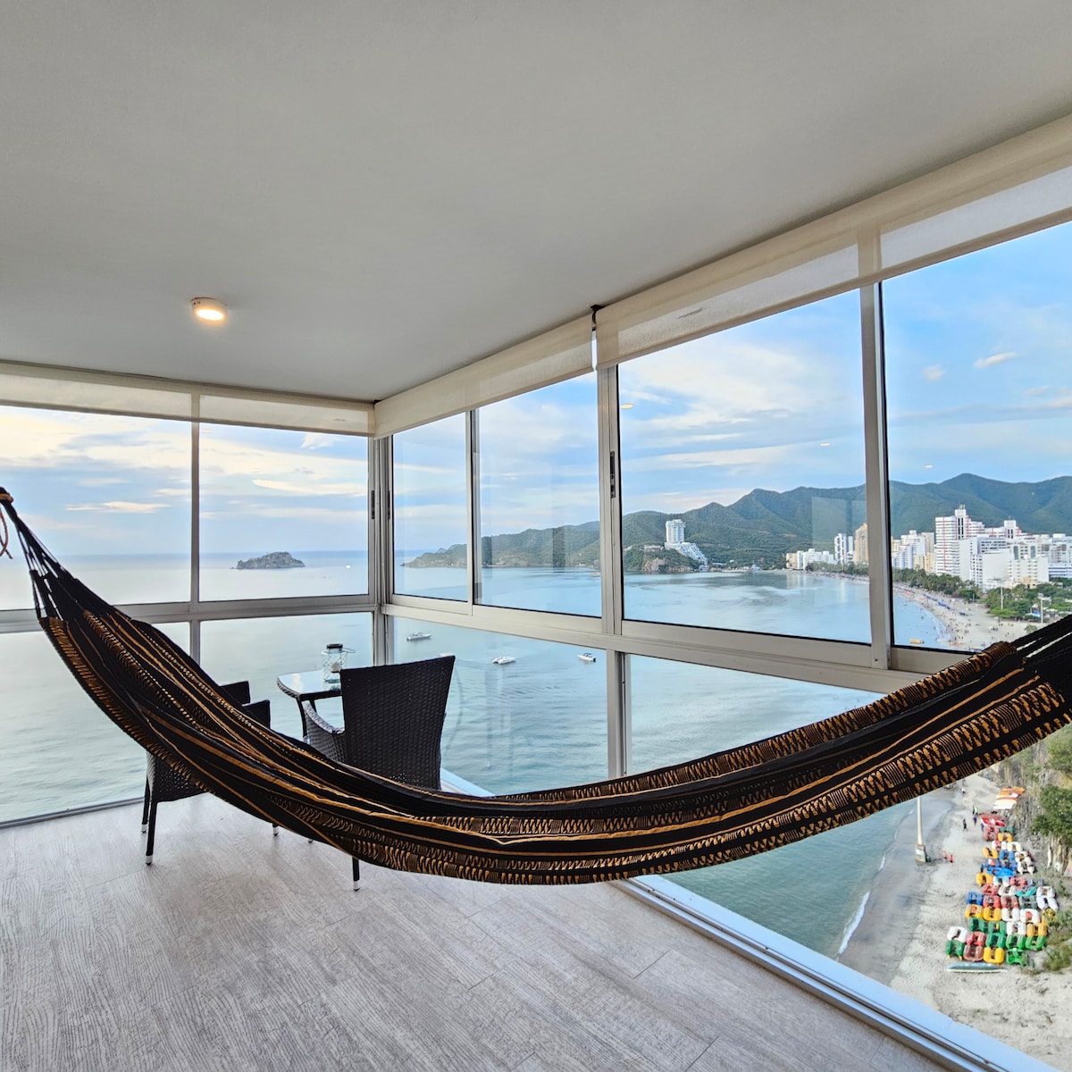 A relaxing corner is highlighted by a hammock with a view of rolling waves and the shoreline. Panoramic windows frame an expansive landscape that includes the beach, mountains, and bustling city life, creating a serene space to unwind.