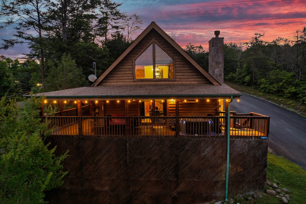 An aerial view of a two-story log cabin showcases a spacious wrap-around deck adorned with ambient lighting. The exterior features triangular gables and large windows, with a wooded backdrop and dusk sky reflecting warm colors.