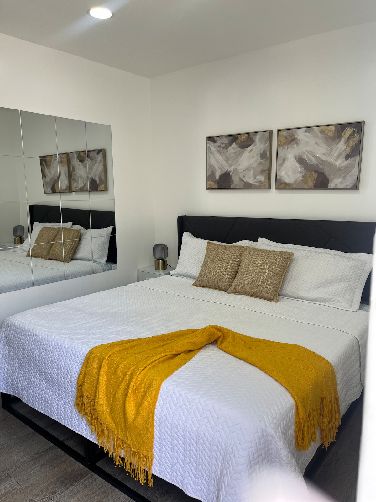 A spacious bedroom features a king-size bed with white bedding and decorative pillows. A bright yellow throw blanket adds a pop of color. Two modern artworks are displayed above the bed, while a large mirror reflects the room's design, enhancing the sense of space.