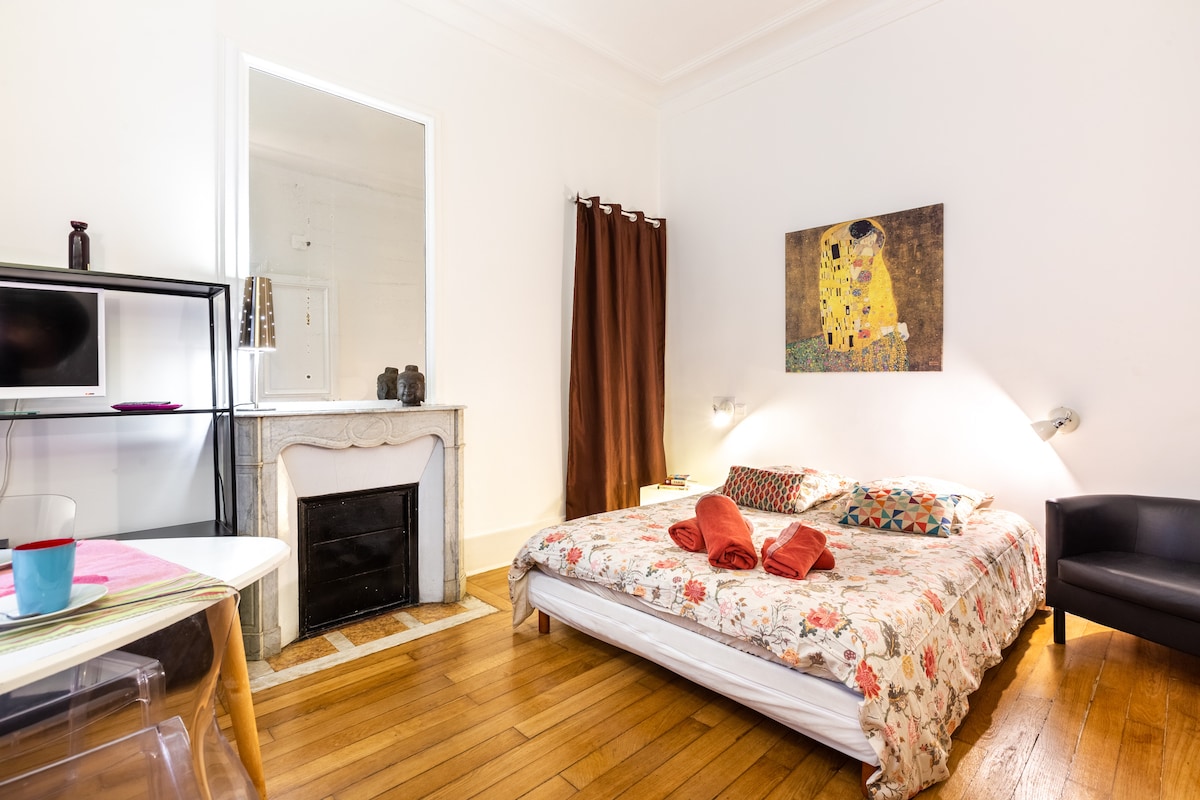 A spacious sleeping area features a floral-patterned bedspread laid over a large bed. A simple chair sits nearby, and a framed artwork is mounted on the wall above. Natural light illuminates the wooden floor, creating a warm ambiance.