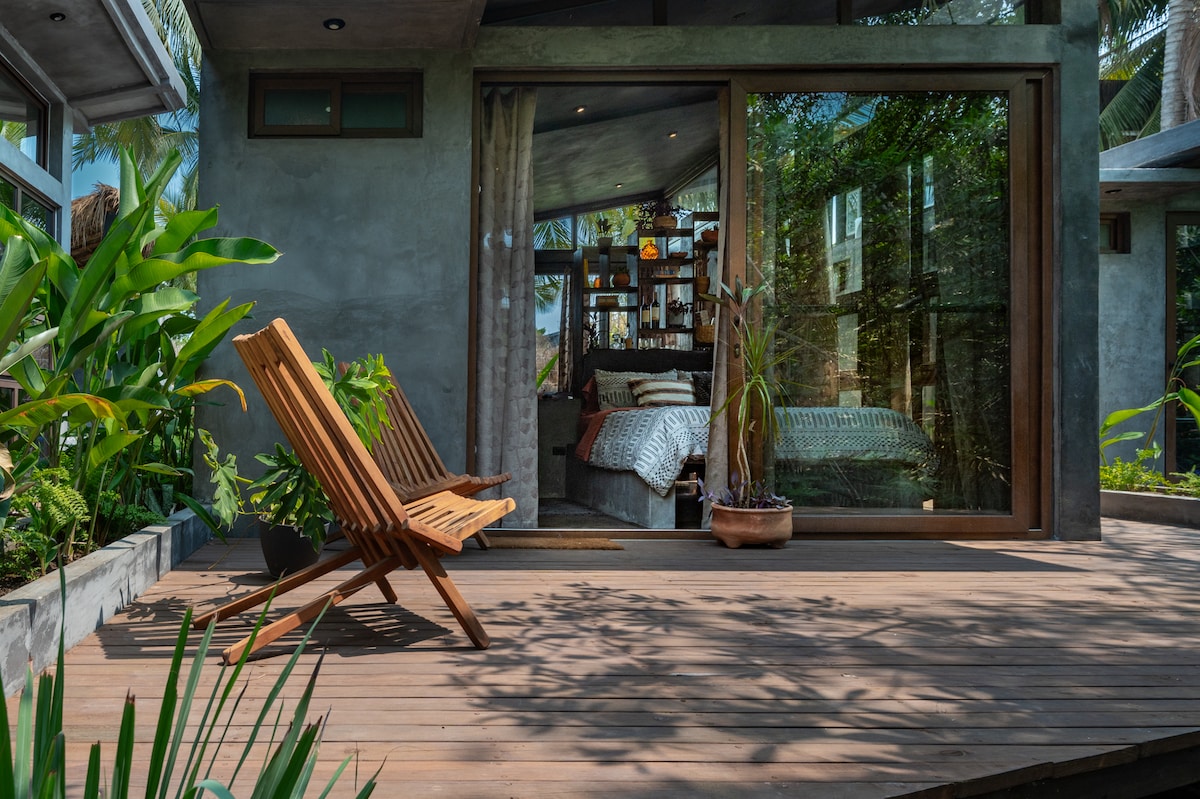 An outdoor wooden deck features two slatted chairs facing large glass doors that provide a view of a cozy interior. Lush greenery surrounds the area, enhancing the connection between indoor and outdoor spaces.