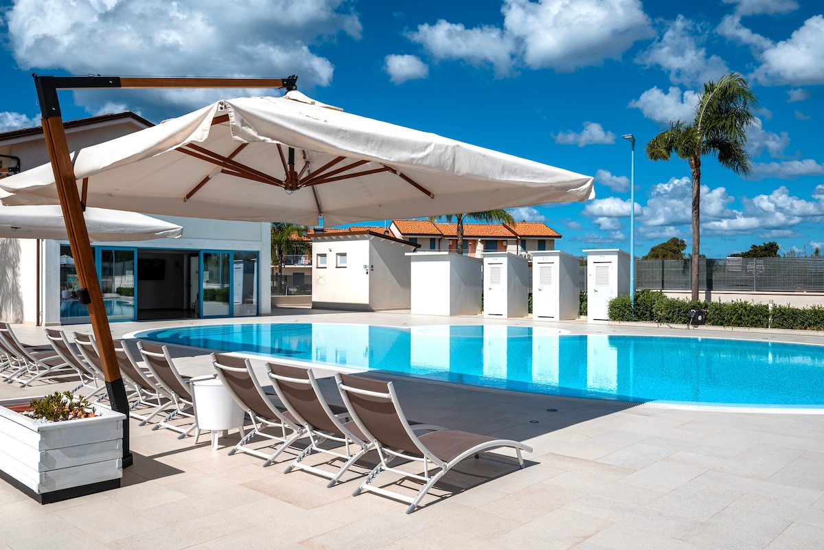 A serene pool area showcases a sparkling water feature surrounded by sun loungers and large shade umbrellas. Palm trees sway gently in the background under a bright blue sky, while modern poolside facilities are visible, enhancing the inviting atmosphere.