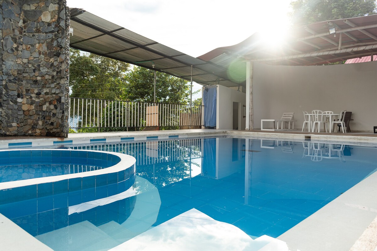 A serene swimming pool is framed by a sunlit patio, showcasing a bright blue surface that reflects the surrounding greenery. Adjacent to the pool, a covered outdoor area features several white chairs and tables, providing space for relaxation and gatherings.