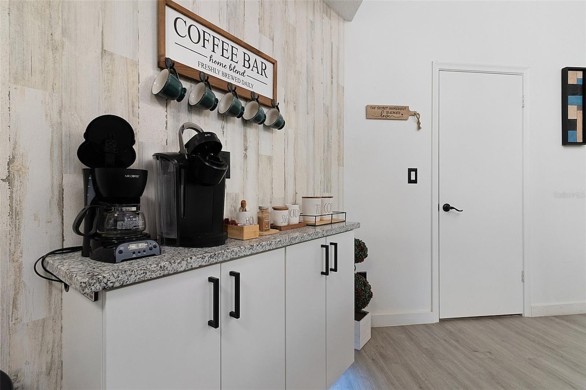 A coffee bar area is showcased with a countertop featuring a coffee maker and a single-serve machine. Above, a sign labeled 'COFFEE BAR' is displayed, complemented by several coffee mugs hanging from hooks. The cabinetry is finished in white, providing a clean look.
