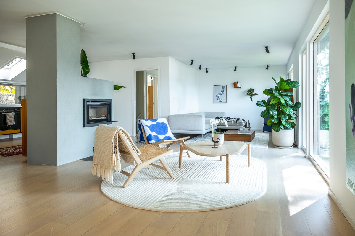 A spacious living area features a comfortable sofa arranged near a cozy fireplace. A light-colored area rug and a round coffee table harmonize with the room's natural elements, including a large potted plant. Bright light filters in through expansive glass doors.